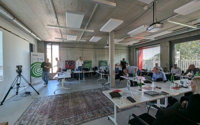 Im Raum sitzen mehrere Personen an Tischen mit Laptops und Unterlagen. In der Ecke steht ein Präsentationsbildschirm. Einige Teilnehmer stehen und diskutieren. Der Raum hat große Fenster, durch die Tageslicht hereinströmt. Ein Teppich liegt auf dem Boden.