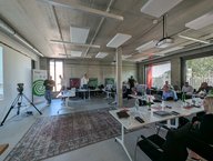 Im Raum sitzen mehrere Personen an Tischen mit Laptops und Unterlagen. In der Ecke steht ein Präsentationsbildschirm. Einige Teilnehmer stehen und diskutieren. Der Raum hat große Fenster, durch die Tageslicht hereinströmt. Ein Teppich liegt auf dem Boden.