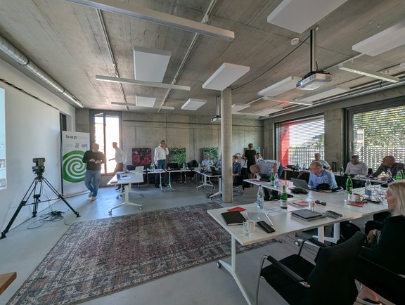 Im Raum sitzen mehrere Personen an Tischen mit Laptops und Unterlagen. In der Ecke steht ein Präsentationsbildschirm. Einige Teilnehmer stehen und diskutieren. Der Raum hat große Fenster, durch die Tageslicht hereinströmt. Ein Teppich liegt auf dem Boden.