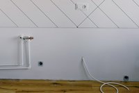 Das Bild zeigt einen Teil einer Wand mit weißen Rohren und einer Wandsteckdose. Die Wand hat diagonale Linien und ist oberhalb des Fußbodens sichtbar. Auf dem Boden verläuft ein längeres, weißes Kabel. Das Holzparkett ist sichtbar und gibt dem Raum eine warme Ausstrahlung.