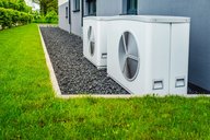 Zwei weiße Klimaanlagen stehen auf einem Kiesbett neben einem modernen Gebäude. Der Rasen ist grün und gepflegt. Die Anlage ist Teil der Außenanlage und bietet Kühlung für das Gebäude.
