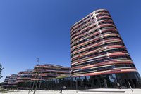 Moderne Gebäudestruktur mit geschwungenem Design und bunten Fassaden in verschiedenen Farben. Im Vordergrund sind Bäume und einige Fußgänger zu sehen. Der klare blaue Himmel bietet einen kontrastierenden Hintergrund zur Architektur.