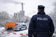 Ein Polizist steht mit dem Rücken zur Kamera in einer verschneiten Umgebung. Im Hintergrund sind ein orangefarbenes Einsatzfahrzeug und ein Polizeiauto mit eingeschaltetem Blaulicht zu sehen. Außerdem gibt es ein großes Gebäude, aus dessen Schornstein Rauch aufsteigt.