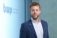 Ein Mann mit einem kurzen, lockigen Bart trägt einen blauen Blazer und ein weißes Hemd. Er steht vor einer Wand mit dem Logo des Bundesverbands Wärmepumpe e.V. und wirkt freundlich.