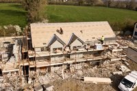 Auf der Baustelle eines Hauses arbeiten mehrere Bauarbeiter. Das Gebäude hat einen unvollendeten Dachstuhl und ist von Gerüsten umgeben. Um das Haus herum liegen Material wie Ziegel und Holz. Im Hintergrund ist eine grüne Wiese zu sehen.