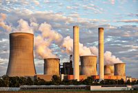 Eine Industrieanlage mit mehreren Kühltürmen und Schornsteinen ist abgebildet. Aus den Türmen und dem Schornstein steigt weißer Dampf auf. Der Himmel ist teils bewölkt und zeigt ein warmes Licht, das die Szene beleuchtet.