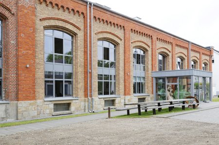Ein historisches Gebäude mit rotem Backstein und großen Fenstern, das modern umgebaut wurde. Im Vordergrund ist ein eingezogener Eingang mit Glas und eine Bank zu sehen. Der Boden ist gepflastert und der Himmel ist bewölkt.