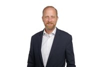 Ein lächelnder Mann mit Bart trägt einen dunkelblauen Anzug und ein weißes Hemd. Er steht vor einem weißen Hintergrund und hat eine entspannte Haltung.