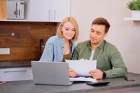 Ein junger Mann und eine junge Frau sitzen an einem Tisch. Der Mann hält Papiere in der Hand, während die Frau ihm zuschaut und lächelt. Auf dem Tisch befindet sich ein Laptop und einige weitere Dokumente. Die Szene vermittelt Zusammenarbeit und Kommunikation.