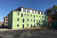 Ein modernes mehrstöckiges Gebäude mit grüner Fassade und weißen Fensterrahmen. Einige Fenster sind ungeschützt. Vor dem Gebäude sind Bauarbeiten im Gange, und es steht ein Bauarbeitsfahrzeug in der Nähe. Der Himmel ist klar und blau.
