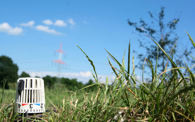 Ein Thermostat steht auf einem Rasen, umgeben von grünem Gras. Im Hintergrund sind Wolken am blauen Himmel und ein hoher Strommast zu sehen. Das Bild vermittelt ein Gefühl von Natur und Technik in einer ländlichen Umgebung.