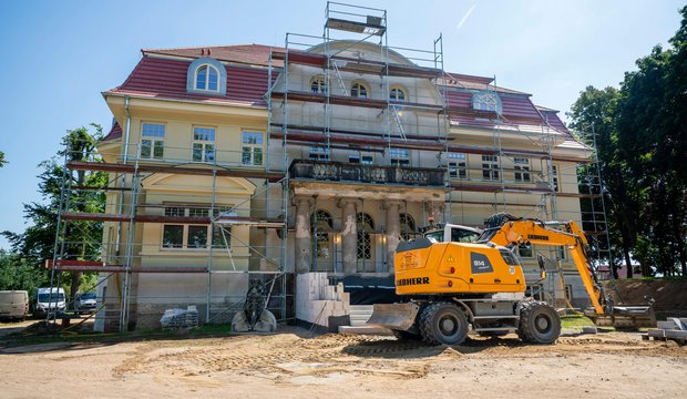 Ein historisches Gebäude wird renoviert. Es ist von Gerüsten umgeben, und ein gelbes Baugerät steht im Vordergrund. Der Boden ist uneben mit sandigem Untergrund, während im Hintergrund Bäume und blauer Himmel sichtbar sind.