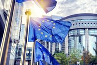 Fünf EU-Flaggen wehen vor einem modernen Gebäude. Die Flaggen sind blau mit gelben Sternen und bewegen sich sanft im Wind. Im Hintergrund sind geometrische Strukturen und Grünpflanzen sichtbar, was auf ein städtisches Umfeld hinweist.