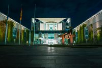 Das Bild zeigt das Kanzleramt in Berlin bei Nacht. Das moderne Gebäude ist von einer hellen Beleuchtung umgeben, und im Vordergrund steht eine große metallische Skulptur. Gru?ne Pflanzen ranken an der Fassade, die einen eindrucksvollen Anblick bietet.