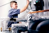 Ein junger Mann in Arbeitskleidung lächelt und arbeitet an einem elektrischen Schaltkasten. Um ihn herum sind verchromte und isolierte Rohre zu sehen. Die Atmosphäre vermittelt eine technische Umgebung, wahrscheinlich in einem Heizungs- oder Klimatechnikbereich.