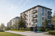 Moderne Wohngebäude mit Balkonen sind von einem gepflegten Park umgeben. Im Vordergrund steht eine große Klimaanlage, die durch digitale Elemente ergänzt wird, die ihre Funktion oder Effizienz symbolisieren. Der Himmel ist klar und die Pflanzen sind grün und lebendig.