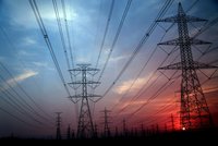 Hochspannungsmasten ragen im Vordergrund gegen einen farbenfrohen Sonnenuntergang auf. Die Masten sind von Stromleitungen umgeben, die sich durch den Himmel ziehen. Der Himmel ist in sanften Blau- und Rosatönen gefärbt.