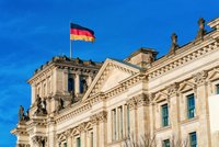 Das Bild zeigt einen Teil des Reichstagsgebäudes in Berlin, das mit der deutschen Flagge geschmückt ist. Der Himmel ist blau, und die beeindruckende Architektur des Gebäudes ist gut sichtbar.