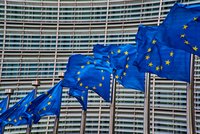 Sechs europäische Flaggen wehen vor einem modernen Gebäude mit einer verspiegelten Fassade. Die Flaggen sind blau mit gelben Sternen und symbolisieren die europäische Einheit. Der Hintergrund zeigt horizontal angeordnete Fensterläden, die das Licht reflektieren.