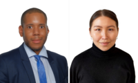 Das Bild zeigt zwei Personen in formeller Kleidung vor einem weißen Hintergrund. Links steht ein Mann mit kurzen Haaren, der einen Anzug mit blauem Hemd und Krawatte trägt. Rechts steht eine Frau mit langen, glatten Haaren in einem schwarzen Rollkragenpullover. Beide sehen in die Kamera.