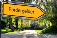 Ein gelbes Schild mit dem Hinweis "Fördergelder" zeigt in eine Richtung. Der Hintergrund zeigt eine grüne Parklandschaft mit Bäumen und Wegen.