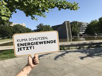 Eine Hand hält ein Schild mit der Aufschrift „KLIMA SCHÜTZEN, ENERGIEWENDE JETZT!“ im Freien. Im Hintergrund sind Bäume und Gebäude zu sehen sowie ein Wasserbecken. Es ist ein sonniger Tag.