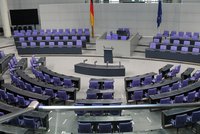 Bildbeschreibung: Der Plenarsaal eines Parlaments mit halbkreisförmigen Sitzen in lila und schwarzen Tischen. In der Mitte befindet sich ein Rednerpult. Hinter dem Pult sind deutsche und europäische Flaggen. Der Raum ist hell und modern gestaltet.