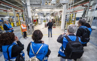 Eine Gruppe von Personen in blauen Westen steht in einer Fabrikhalle. Ein Mann in schwarzer Kleidung spricht zu ihnen, während einige der Anwesenden Notizen machen oder Fotos mit ihren Handys aufnehmen. Im Hintergrund sind industrielle Maschinen und Ausrüstungen zu sehen.