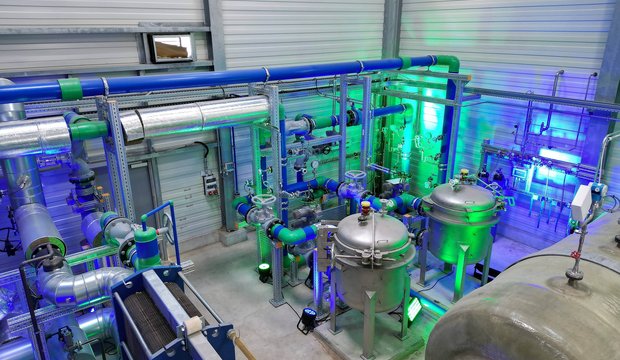 In der Abbildung ist eine industrielle Anlage mit Rohren, Tanks und Ventilen zu sehen. Grüne und blaue Lichter beleuchten die Ausrüstung. Die Struktur deutet auf einen technischen Prozess zur Wasser- oder Luftbehandlung hin.