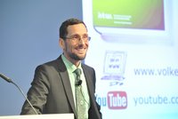 Ein lächelnder Mann in einem Anzug steht vor einem Podium. Er trägt ein mintgrünes Hemd und eine Krawatte mit Muster. Im Hintergrund sind mehrere Webseiten und Logos sichtbar. Der Mann scheint zu präsentieren und spricht vielleicht zu einem Publikum.