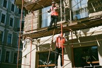 Zwei Bauarbeiter stehen auf einem Gerüst vor einem Gebäude. Einer der Arbeiter nutzt ein Werkzeug, während der andere auf der oberen Ebene steht. Beide tragen Sicherheitswesten. Das Gerüst ist am Gebäude angebracht und bei Tageslicht aufgenommen.