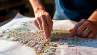 Eine Hand zeigt auf eine detaillierte Stadtkarte, die in 3D dargestellt ist. Die Karte zeigt verschiedene Gebäude und Straßen. Die Person scheint einen bestimmten Punkt oder Bereich auf der Karte zu markieren oder zu erkunden.
