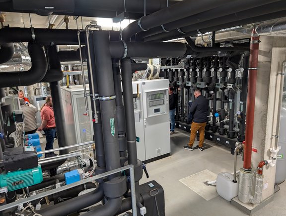 In einem technischen Raum sind verschiedene Heizungs- und Lüftungsanlagen zu sehen. Schwarz-rohre und Maschinen sind sichtbar, während eine Person an einem Bedienfeld arbeitet. Eine andere Person ist im Hintergrund zu sehen. Der Raum wirkt gut strukturiert und organisiert.