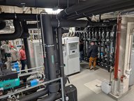 In einem technischen Raum sind verschiedene Heizungs- und Lüftungsanlagen zu sehen. Schwarz-rohre und Maschinen sind sichtbar, während eine Person an einem Bedienfeld arbeitet. Eine andere Person ist im Hintergrund zu sehen. Der Raum wirkt gut strukturiert und organisiert.