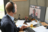 Ein Mann mit Headset sitzt vor einem Computer, auf dem eine Videokonferenz läuft. Er betrachtet den Bildschirm, auf dem eine Person spricht. Auf dem Schreibtisch sind Unterlagen und ein Getränk sichtbar. Der Raum hat große Fenster mit Blick nach draußen.