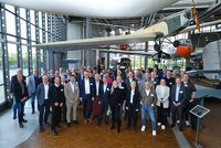 Eine Gruppe von über 40 Personen steht in einem großen Raum mit Flugzeugen und historischen Exponaten im Hintergrund. Die Menschen lächeln und tragen überwiegend formelle Kleidung, einige haben Namensschilder an. Der Raum hat große Fenster, die Tageslicht hereinlassen.
