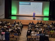 Eine Person steht auf einer Bühne und spricht zu einem Publikum. Im Hintergrund ist eine Präsentation mit einem Europa-Standortplan und Fragen zur Klimaneutralität 2045 sichtbar. Das Publikum besteht aus vielen Menschen, die aufmerksam zuhören.