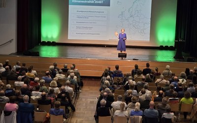 Eine Person steht auf einer Bühne und spricht zu einem Publikum. Im Hintergrund ist eine Präsentation mit einem Europa-Standortplan und Fragen zur Klimaneutralität 2045 sichtbar. Das Publikum besteht aus vielen Menschen, die aufmerksam zuhören.