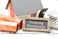 Ein Modellhaus steht neben Kupferrohren und einem Heizkörperthermostat. Ein Schild mit der Aufschrift „Förderung Wärmepumpe“ liegt prominent im Vordergrund. Die Bildkomposition thematisiert Heiztechnik und mögliche Förderungen für Wärmepumpen.