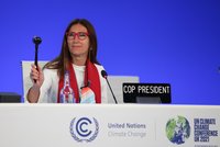 Eine Frau mit langen Haaren, die eine rote Brille und einen roten Schal trägt, sitzt an einem Tisch. Sie hält einen Hammer in der Hand und lächelt. Auf dem Tisch steht das Schild „COP PRESIDENT“ und weitere Informationen zur UN-Klimakonferenz. Der Hintergrund ist blau.