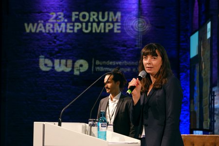 Eine Frau spricht am Rednerpult bei der 23. Forum Wärmepumpe, unterstützt von einem Mann im Hintergrund. Die Bühne ist beleuchtet, und das Thema der Veranstaltung ist auf einer Leinwand sichtbar. Es herrscht eine professionelle Atmosphäre.