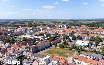 Luftaufnahme einer Stadt mit roten Dächern, Wohngebäuden und offenen Feldern im Hintergrund. Die Stadt zeigt eine Mischung aus modernen und traditionellen Architekturen, umgeben von natürlicher Landschaft und blauem Himmel mit einigen Wolken.