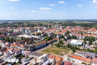 Luftaufnahme einer Stadt mit roten Dächern, Wohngebäuden und offenen Feldern im Hintergrund. Die Stadt zeigt eine Mischung aus modernen und traditionellen Architekturen, umgeben von natürlicher Landschaft und blauem Himmel mit einigen Wolken.