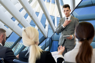 Ein junger Mann in einem Anzug präsentiert in einem modernen, lichtdurchfluteten Raum vor drei Zuhörern. Er hält ein Tablet in der Hand und gestikuliert, um seine Punkte zu verdeutlichen. Die Zuhörer sind aufmerksam und blicken in seine Richtung.