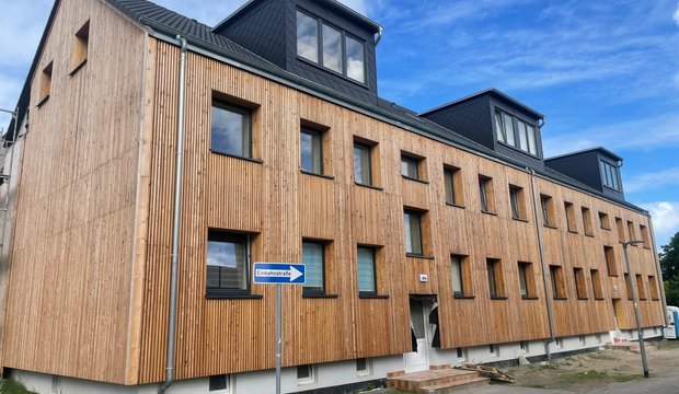 Ein modernes Gebäude mit Holzverkleidung und schwarzen Dachelementen. Es hat mehrere Fenster und Eingänge, die gleichmäßig angeordnet sind. Im Vordergrund steht ein Verkehrsschild, das auf eine Einbahnstraße hinweist. Der Himmel ist blau mit einigen Wolken.