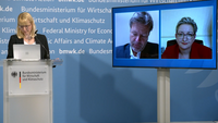 Eine Person steht an einem Podium mit dem Logo des Bundesministeriums für Wirtschaft und Klimaschutz. Auf zwei Bildschirmen sind weitere Personen zu sehen, die an einer virtuellen Konferenz teilnehmen. Der Hintergrund ist hellblau mit Schriftzügen des Ministeriums.