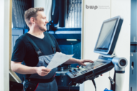 Ein lächelnder Techniker in Arbeitskleidung steht an einer Maschinensteuerung und hält einen Plan in der Hand. Er fokussiert auf den Bildschirm der Maschine, während im Hintergrund eine industrielle Umgebung sichtbar ist.