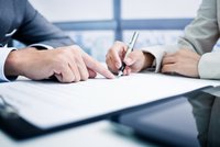 Zwei Hände sind über einem Dokument positioniert. Eine Hand zeigt auf einen Punkt auf dem Papier, während die andere Hand mit einem Stift das Dokument bearbeitet. Es handelt sich um eine Gesprächssituation, vermutlich zur Vertragsunterzeichnung oder Dokumentenüberprüfung.