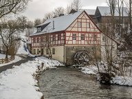 Ein historisches, zweigeschossiges Gebäude mit Fachwerkfassade steht an einem schneebedeckten Ufer. Neben dem Haus fließt ein ruhiger Wasserlauf, in dem ein Wasserrad sichtbar ist. Die Umgebung ist winterlich mit schneebedeckten Bäumen und einem grauen Himmel.