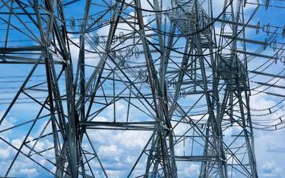 Ein Blick auf Hochspannungsmasten, die in den Himmel ragen. Die Struktur ist aus metallischen Elementen gefertigt, umgeben von einem blauen Himmel mit vereinzelten Wolken. Die Masten sind Teil des Stromnetzes und transportieren elektrische Energie.